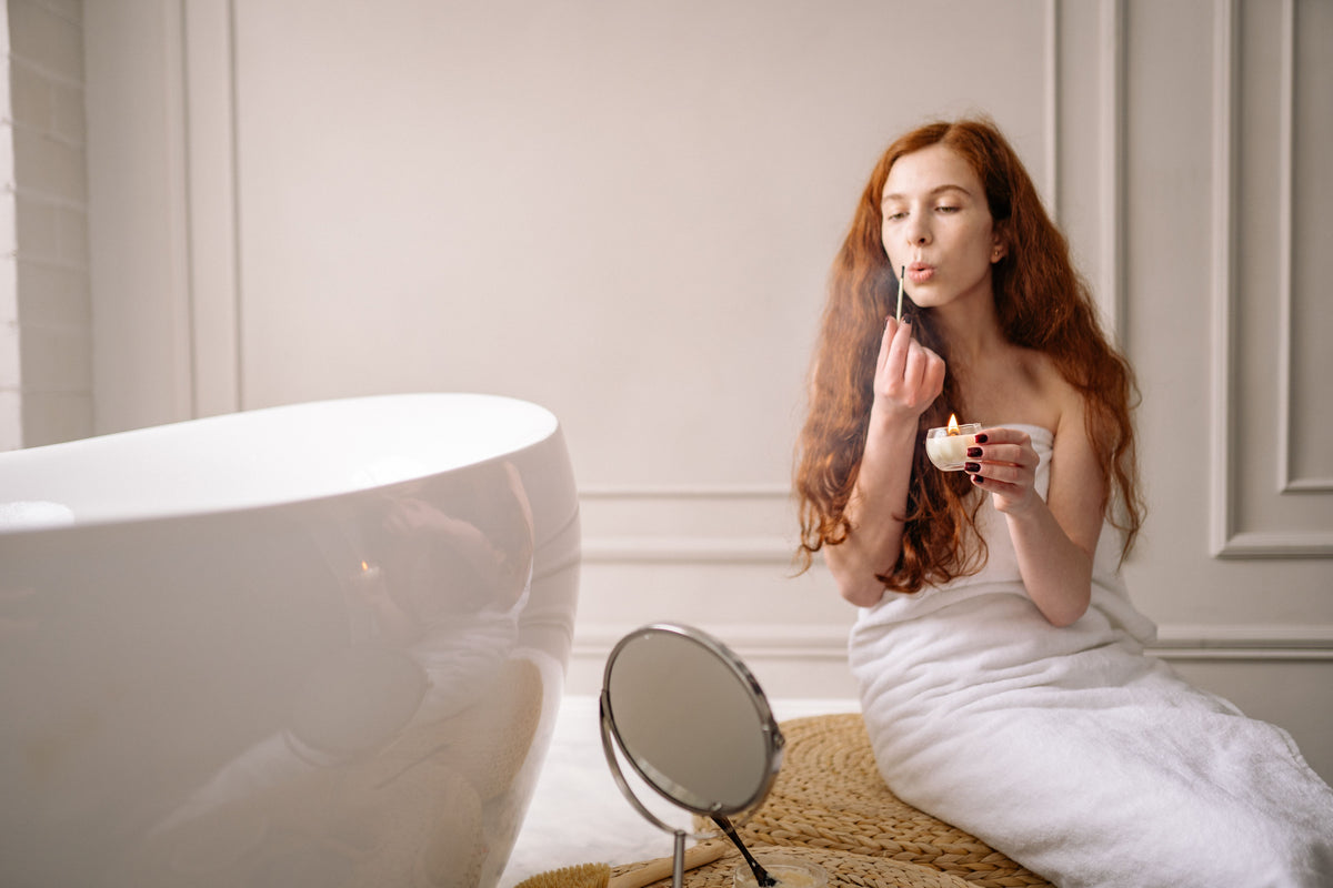 Red-haired woman wrapped in towel, lighting a candle in a luxurious bathroom for a relaxing experience.